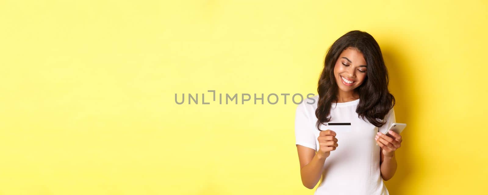 Young attractive woman shopping online, using mobile phone with credit card and smiling, standing over yellow background.