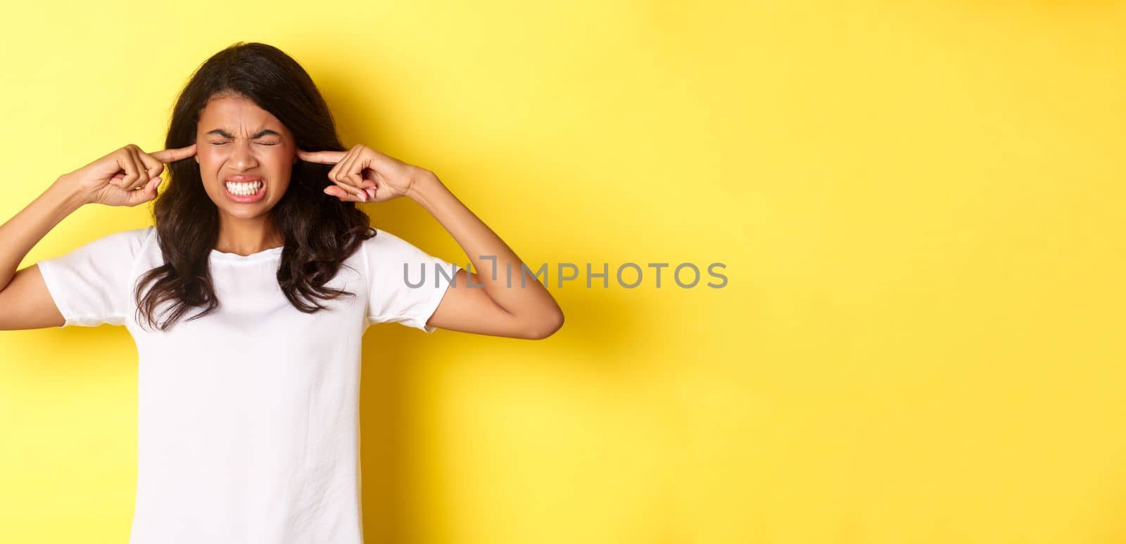Portrait of annoyed and pissed-off african-american girl, shut her ears and grimacing from loud awful noise, being disturbed by something loud, standing over yellow background.