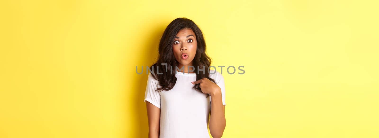 Portrait of surprised african-american girl, pointing finger at herself, standing over yellow background.