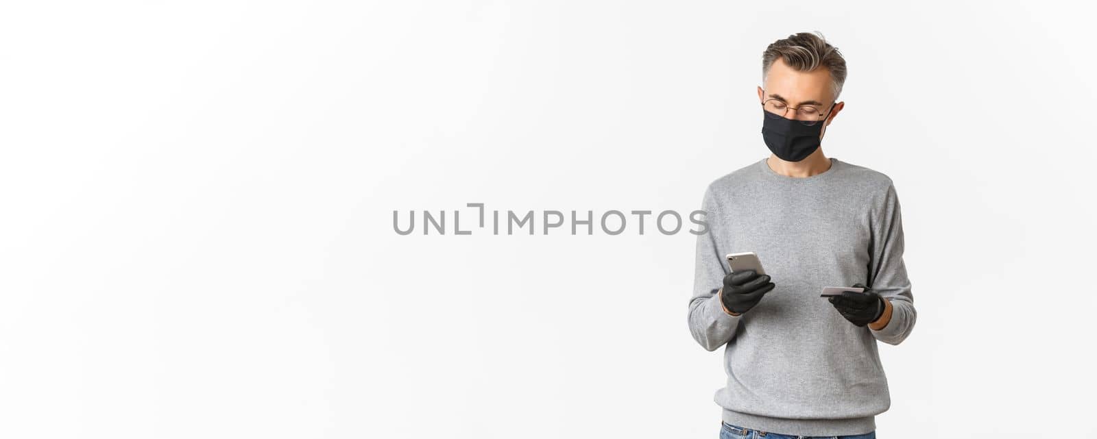 Concept of covid-19, social distancing and lifestyle. Image of handsome middle-aged man in medical mask, gloves and glasses, making online order using credit card and mobile phone by Benzoix