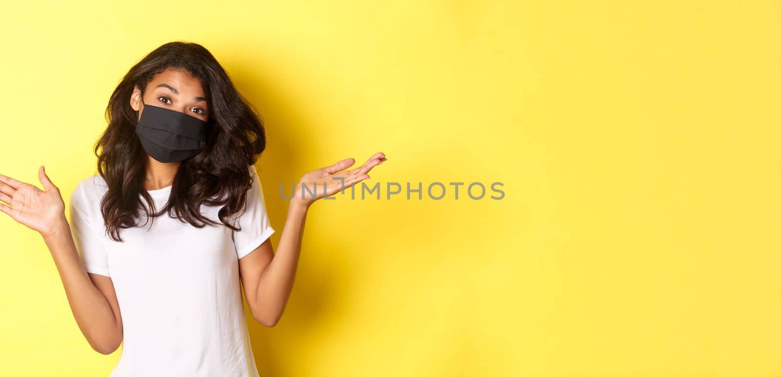 Concept of coronavirus, pandemic and lifestyle. Image of cute african-american girl in face mask, shrugging and looking clueless, dont know anything, standing over yellow background by Benzoix