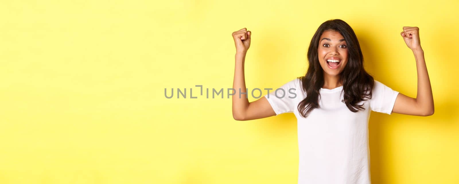 Image of happy african-american girl achieve goal and celebrating victory, raising hands up and smiling pleased, looking satisfied, standing over yellow background by Benzoix