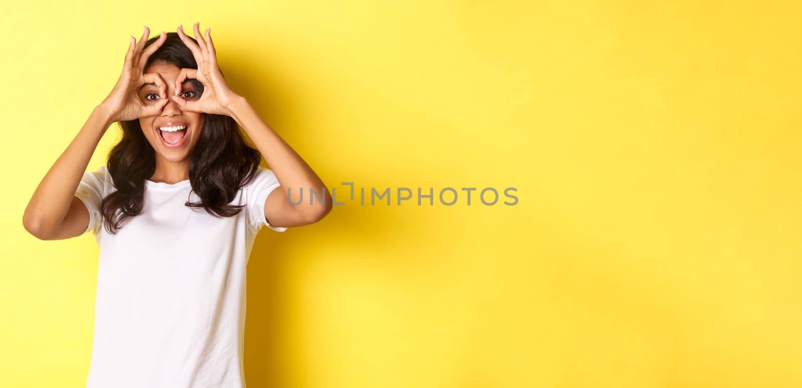 Image of happy and funny african-american girl, making finger glasses and looking through them at something amazing, standing over yellow background.