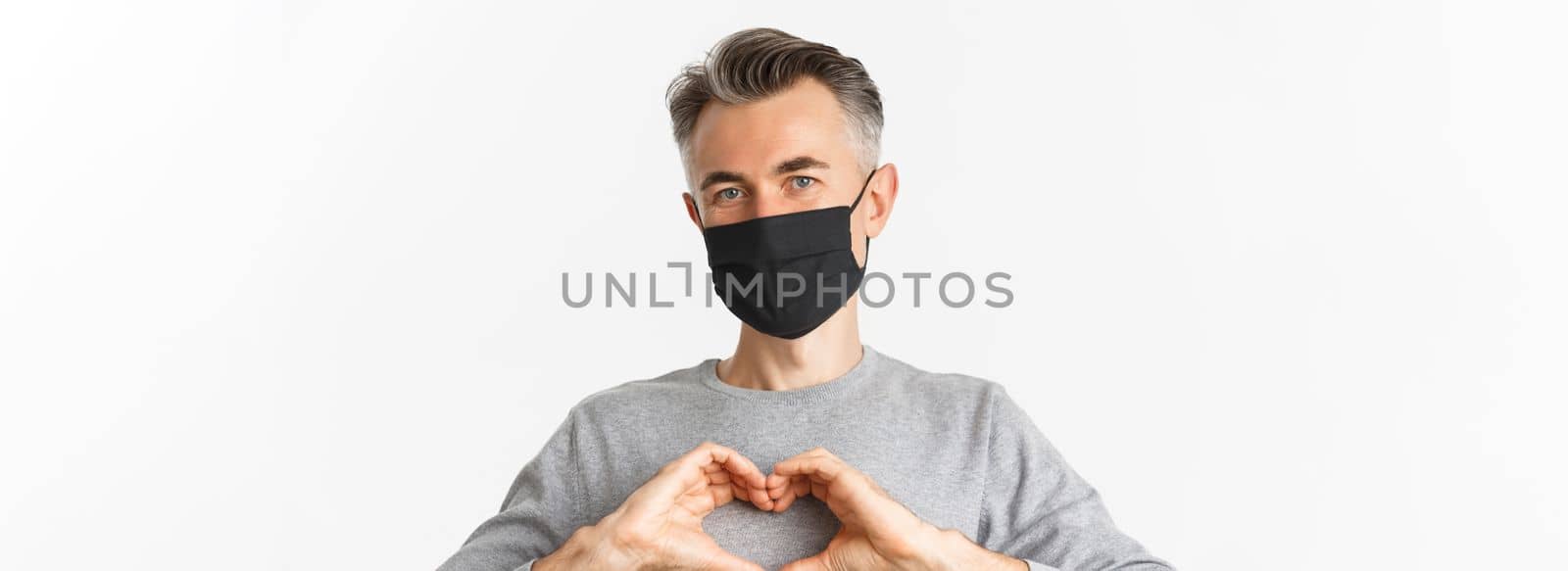 Concept of covid-19, social distancing and quarantine. Close-up of attractive middle-aged man in black medical mask, showing heart sign and smiling, standing over white background.