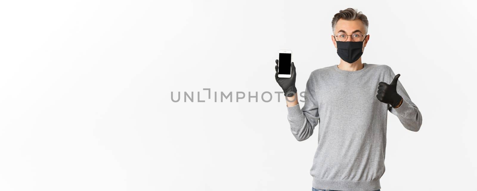 Concept of covid-19, social distancing and lifestyle. Image of satisfied handsome middle-aged man, wearing medical mask, gloves and glasses, showing thumbs-up and mobile screen by Benzoix