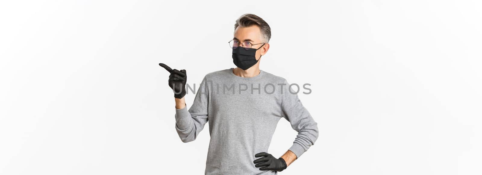 Concept of coronavirus, lifestyle and quarantine. Intrigued middle-aged man in medical mask, gloves and glasses pointing finger right and looking at advertisement, standing over white background.