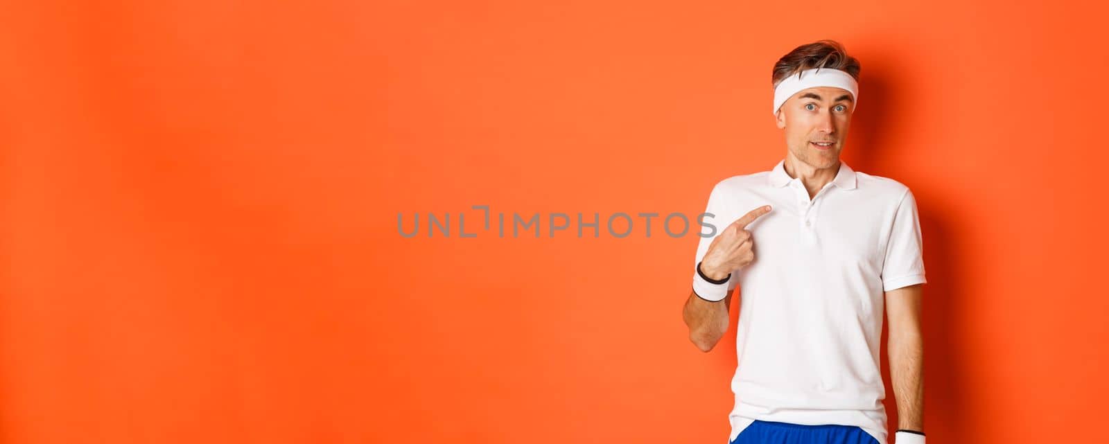 Concept of sport, fitness and lifestyle. Portrait of confused middle-aged sportsman pointing at himself, raising eyebrows puzzled, standing over orange background.