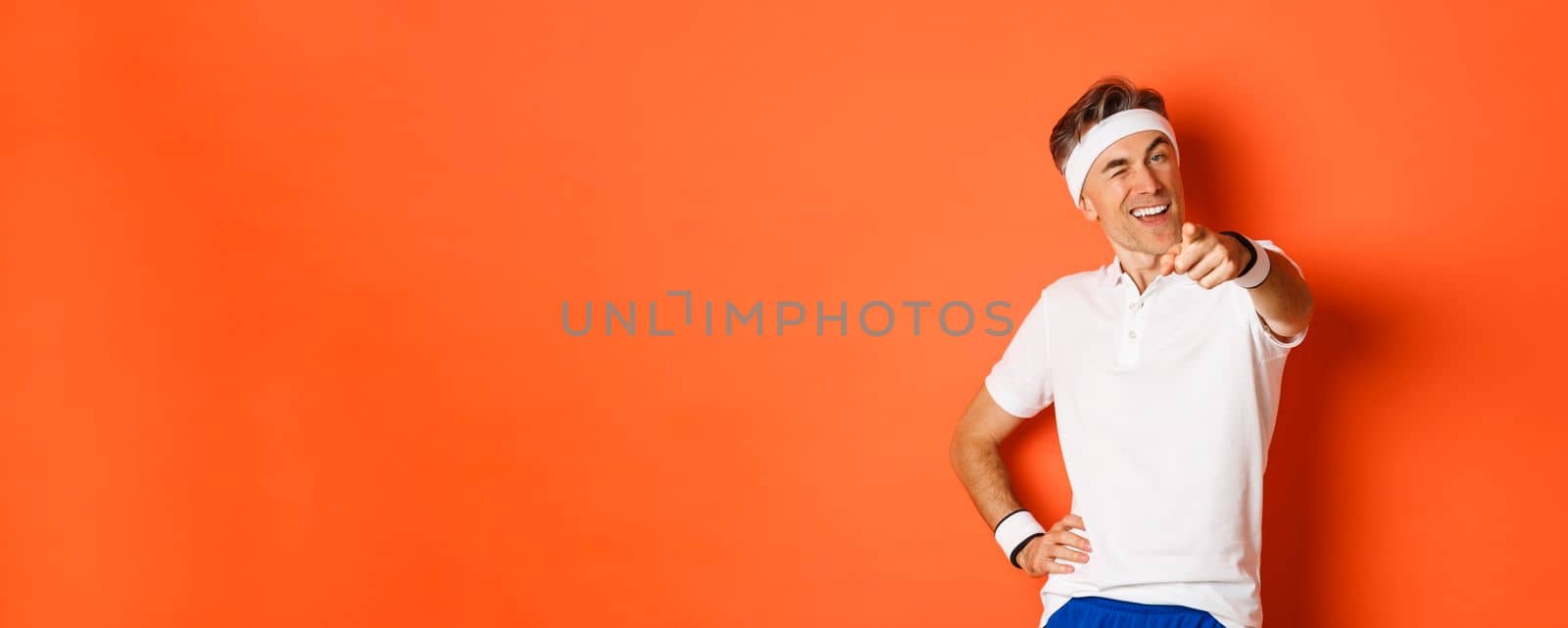 Concept of sport, fitness and lifestyle. Portrait of handsome, healthy middle-aged male athlete, pointing finger at camera and winking, inviting you to gym, standing over orange background.