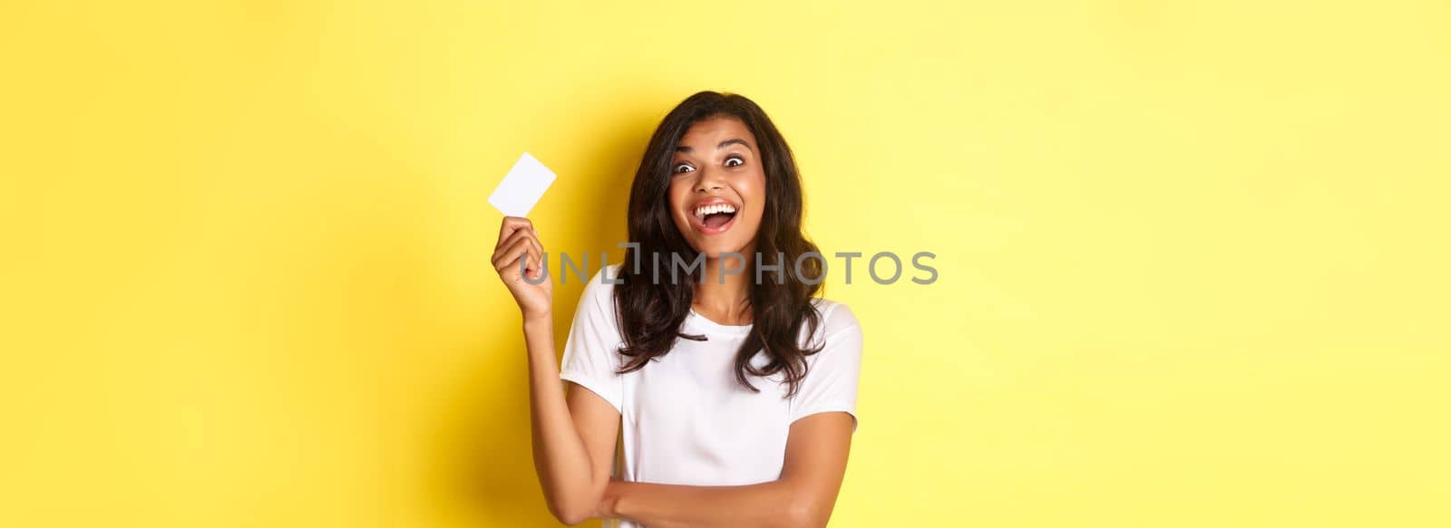 Image of excited african-american woman, showing her credit card and looking amazed at something, going shopping, standing over yellow background.