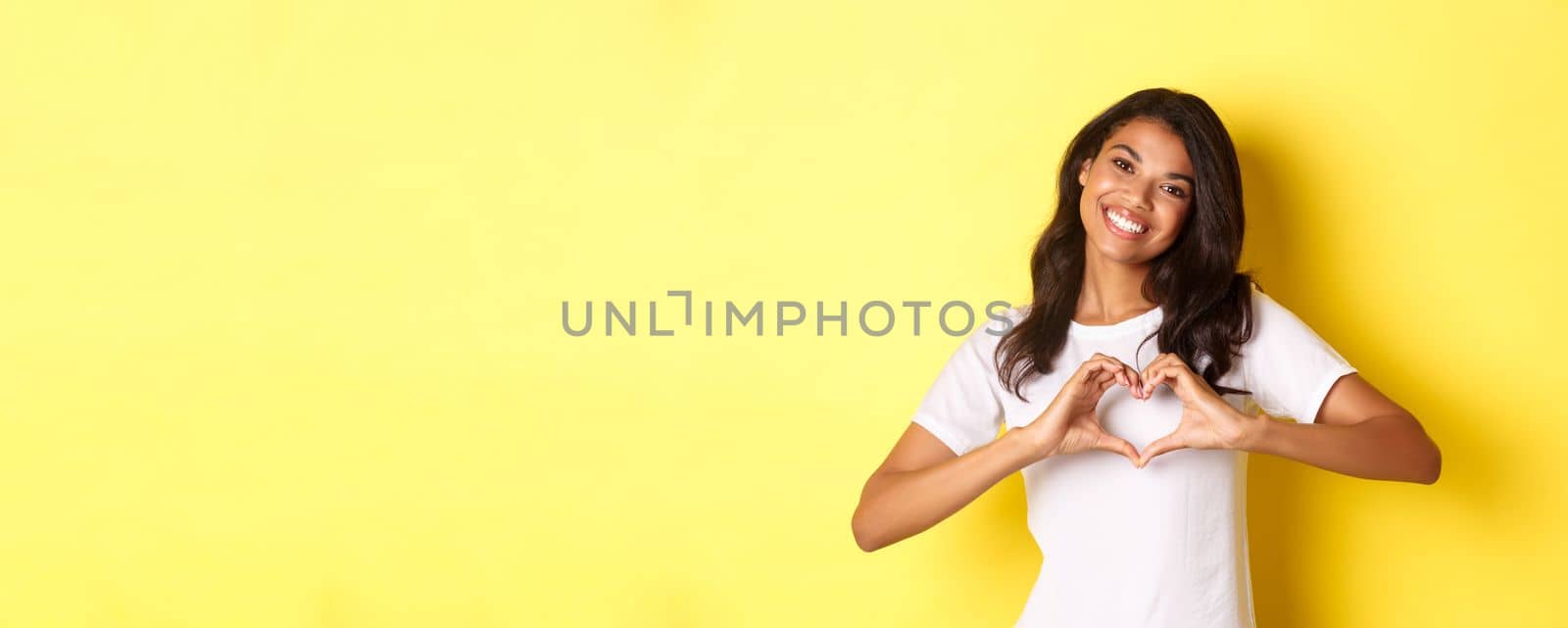 Image of lovely african-american girl in white t-shirt, showing heart sign and smiling, standing over yellow background by Benzoix
