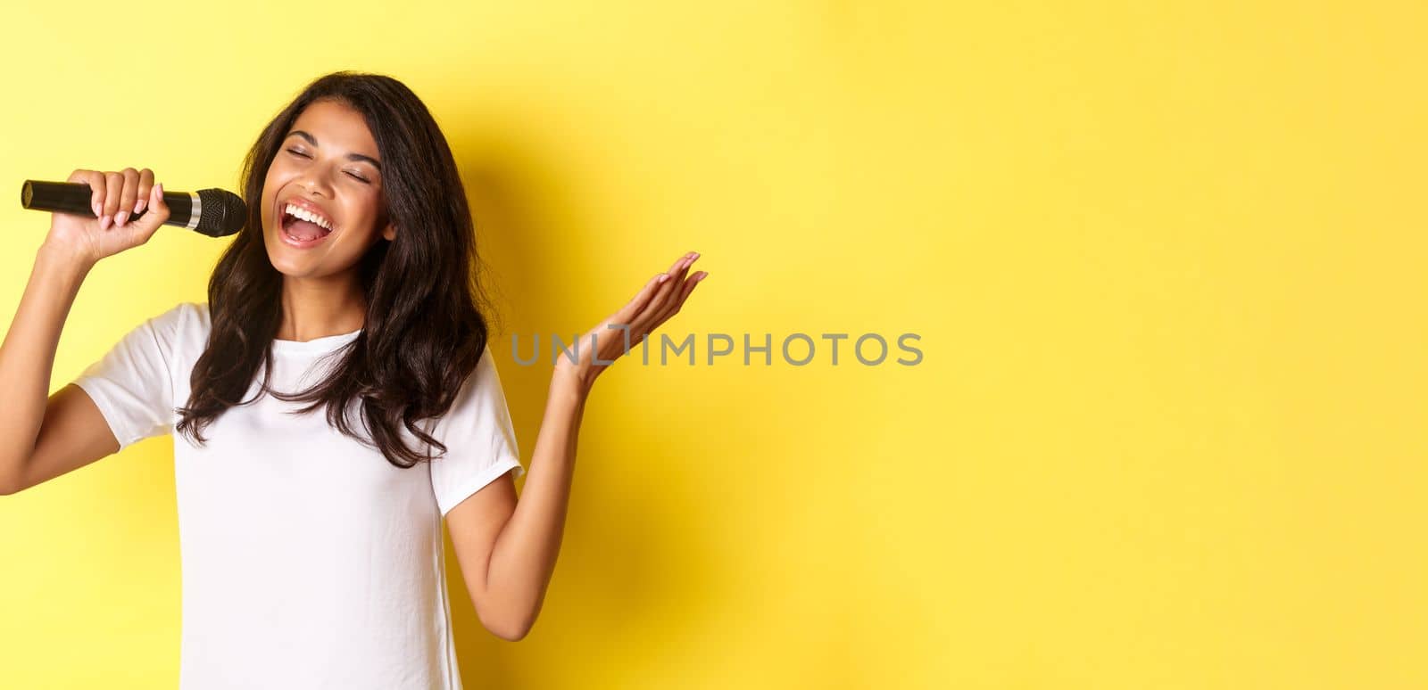 Image of carefree african-american girl singing in microphone, smiling happy, performing over yellow background.