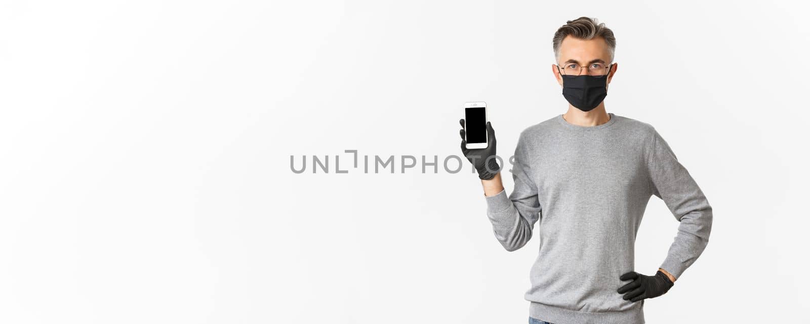 Concept of covid-19, social distancing and lifestyle. Image of confident middle-aged man in glasses, medical mask and gloves, showing smartphone screen, demonstrate an app by Benzoix