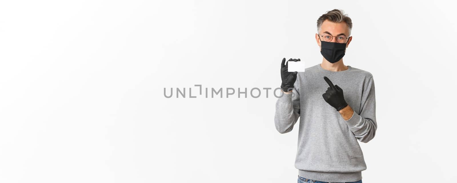Concept of covid-19, social distancing and lifestyle. Handsome middle-aged man in medical mask, gloves and glasses, pointing finger at credit card, standing over white background by Benzoix