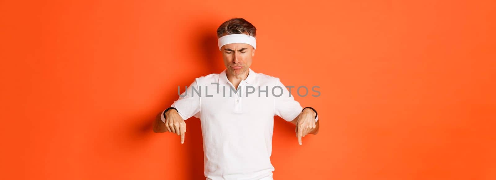 Concept of workout, sports and lifestyle. Image of disappointed and sad adult sportsman, looking and pointing down distressed, frowning upset, standing over orange background.