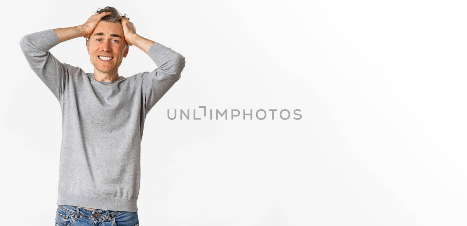 Image of frustrated middle-aged man, holding hands on head and looking distressed, losing something, standing over white background.