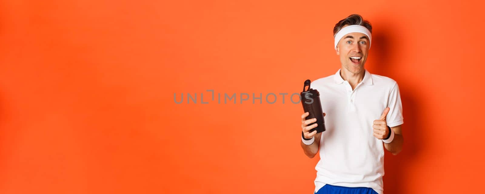 Portrait of healthy and active middle-aged fitness guy, wearing gym uniform, showing thumbs-up, drinking water, standing over orange background.