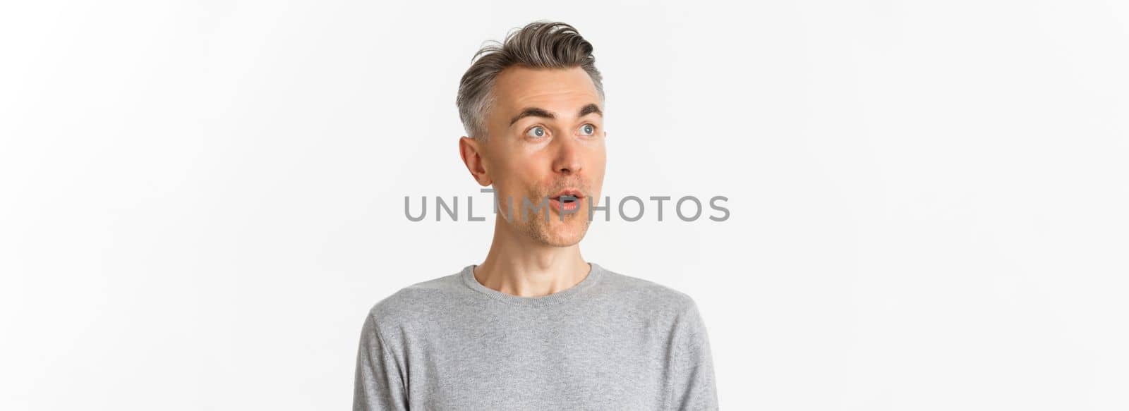Close-up of handsome, middle-aged guy with gray hair, open mouth and looking amazed at upper left corner, standing against white background.