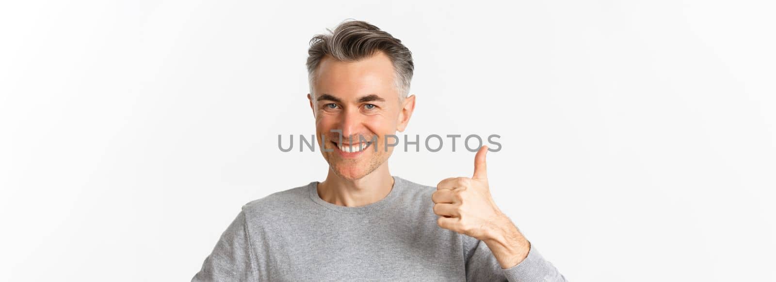 Close-up of handsome, confident middle-aged man in gray sweater, showing thumbs-up in approval, recommending something, standing over white background.