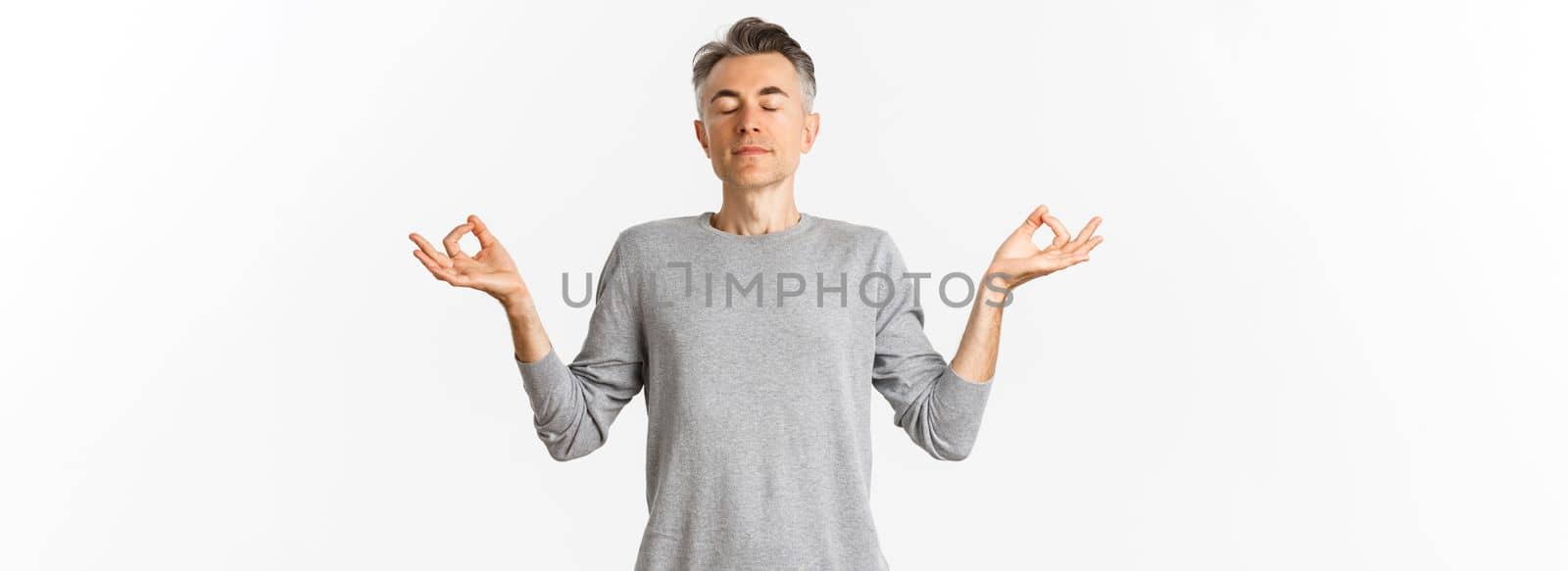 Portrait of calm and relaxed middle-aged man, breathing air freely, meditating with eyes closed and hands spread sideways, practice yoga over white background.