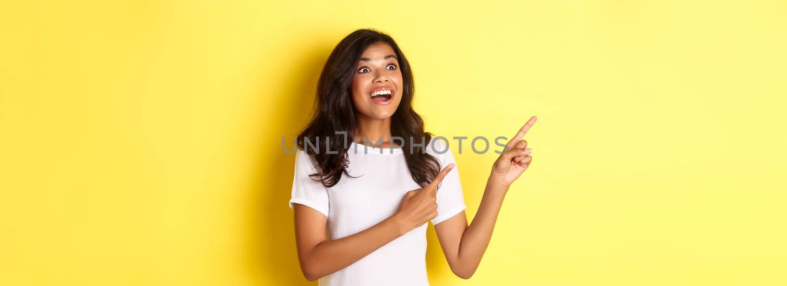 Portrait of beautiful african-american woman, pointing fingers and looking amazed at upper left corner, showing awesome promo offer, standing over yellow background.