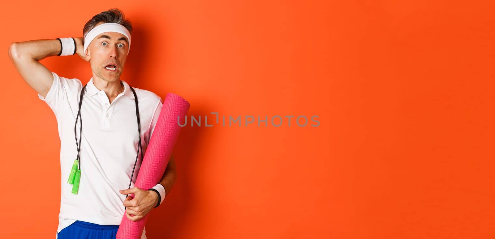 Concept of workout, gym and lifestyle. Image of awkward middle-aged athlete, looking confused, holding yoga mat and skipping rope, standing over orange background.