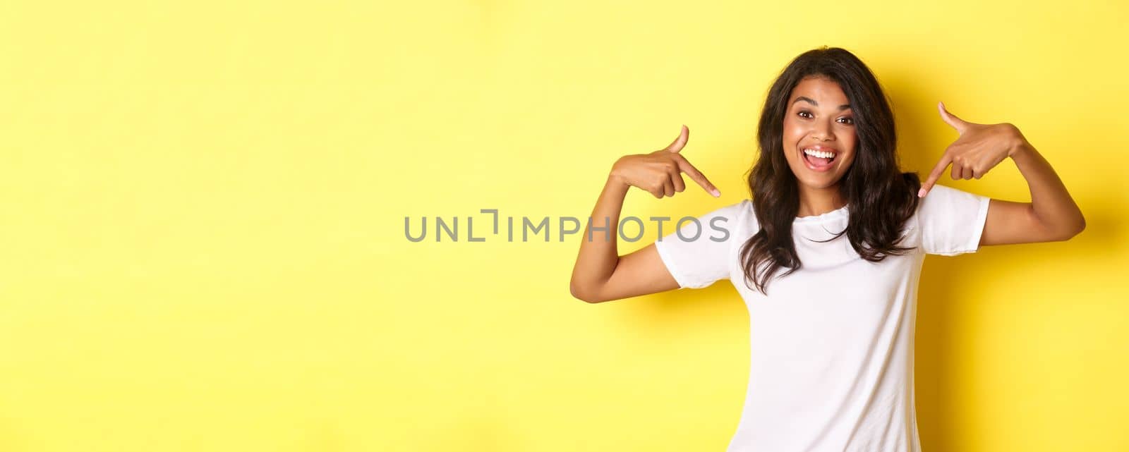 Portrait of cheerful african-american girl in white t-shirt, pointing fingers at your logo on center, standing over yellow background and smiling.