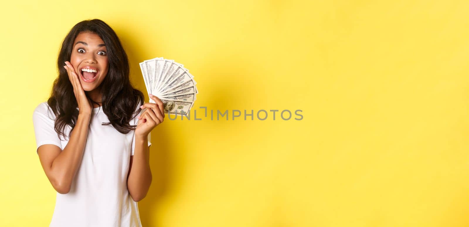 Image of excited lucky girl, winning money and smiling amazed, standing over yellow background.