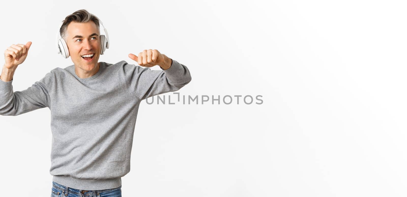 Portrait of handsome, modern middle-aged man, listening music in headphones and dancing, standing over white background.