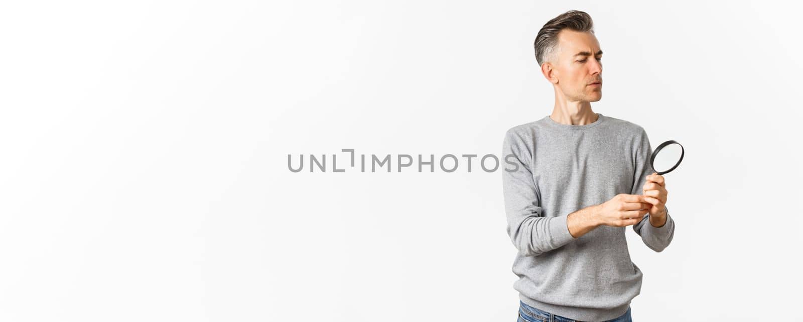 Portrait of thoughtful middle-aged man looking through magnifying glass, searching for something or reading, standing over white background.