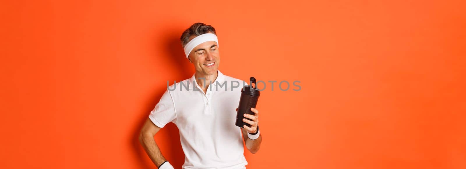 Portrait of pleased middle-aged fitness guy, drinking water and smiling satisfied after productive workout, standing over orange background.