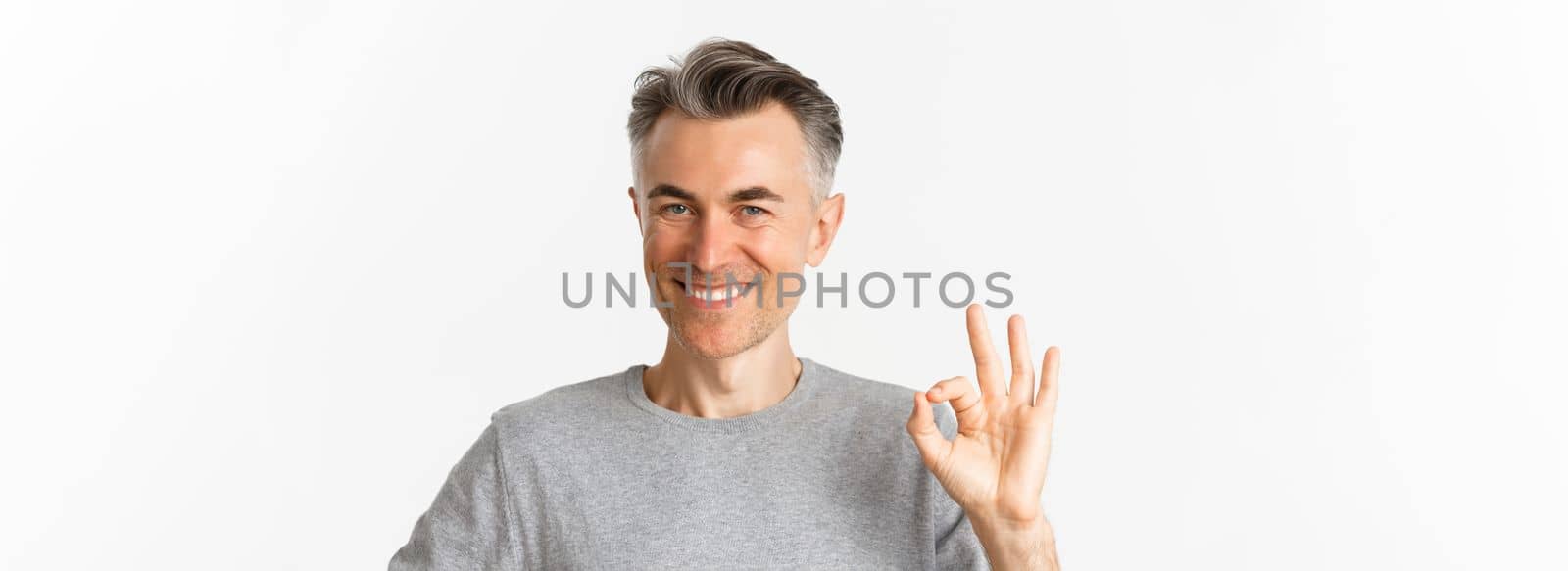 Close-up of handsome middle-aged man, showing okay sign and smiling, guarantee quality, assure that everything good, standing over white background.