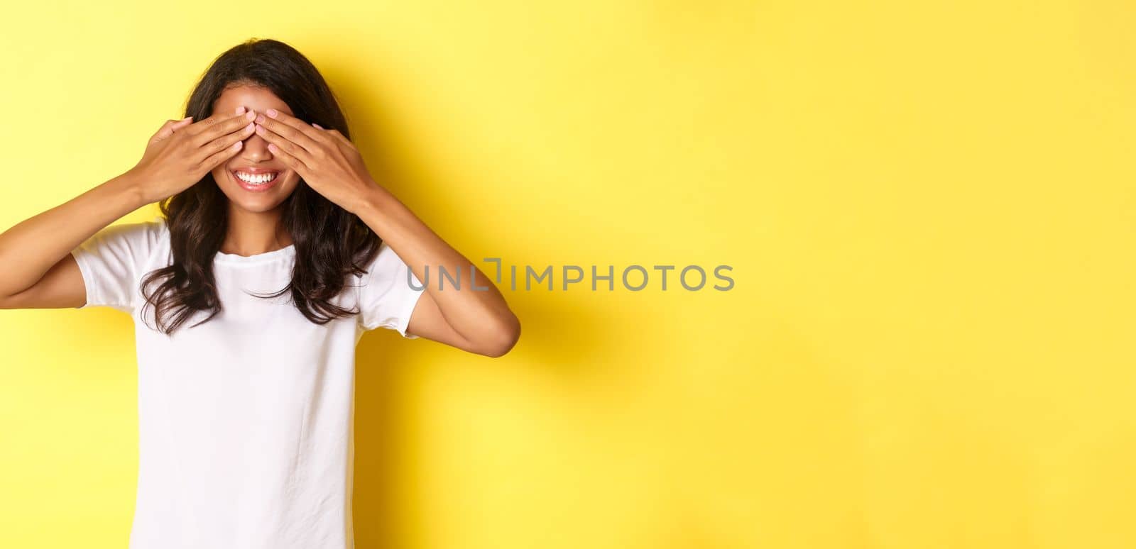 Image of excited african-american girl waiting for surprise, smiling and covering eyes with hands, standing over yellow background.