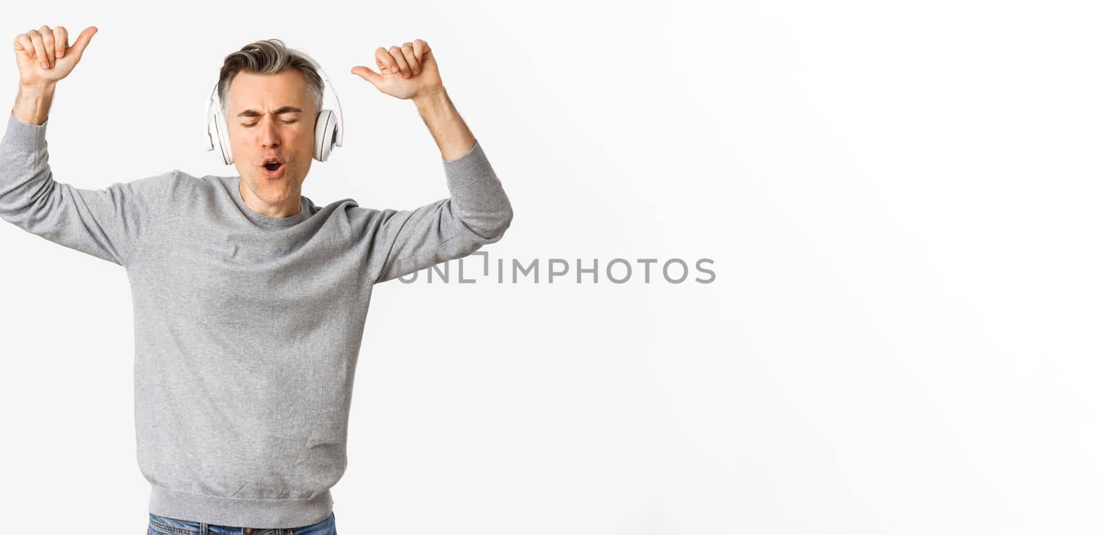 Portrait of handsome, modern middle-aged man, listening music in headphones and dancing, standing over white background.