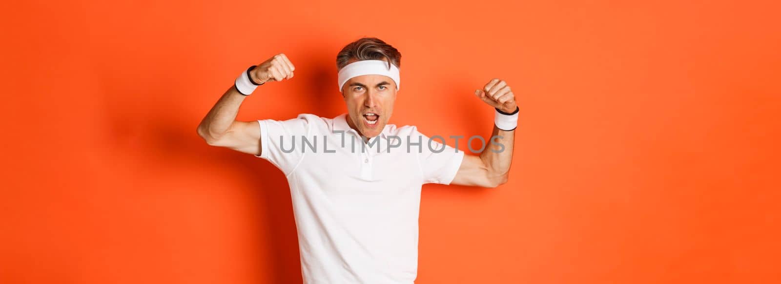 Image of funny middle-aged sportsman in white headband and t-shirt, flexing biceps to show-off, standing against orange background.