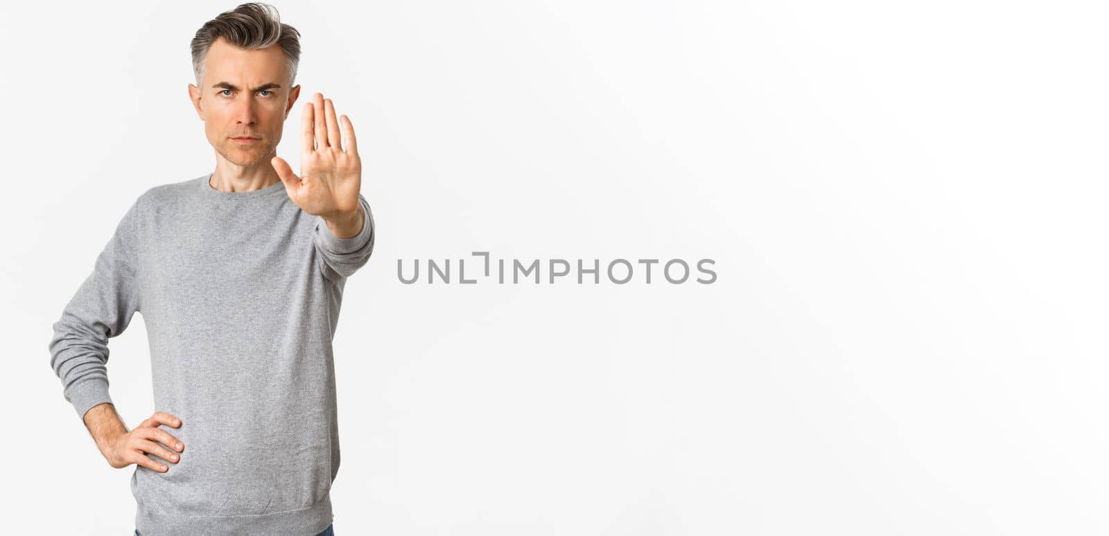 Image of serious middle-aged man extend one hand to stop you, prohibit something bad, frowning and looking with disapproval, standing over white background.