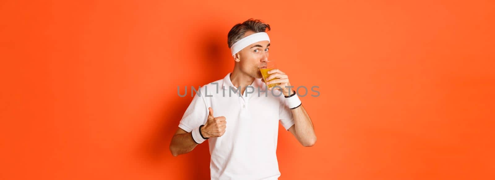 Concept of workout, gym and lifestyle. Portrait of handsome middle-aged fitness guy, showing thumbs-up and drinking juice, standing over orange background.