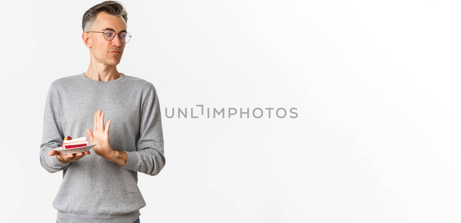 Portrait of displeased middle-aged man, looking away disgusted and rejecting piece of cake, standing bothered over white background.