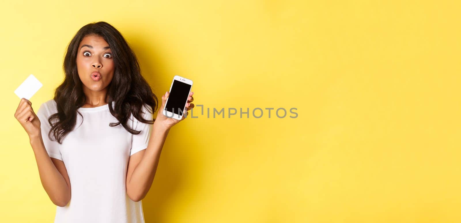 Image of excited african-american female model, showing smartphone screen and credit card, standing over yellow background.