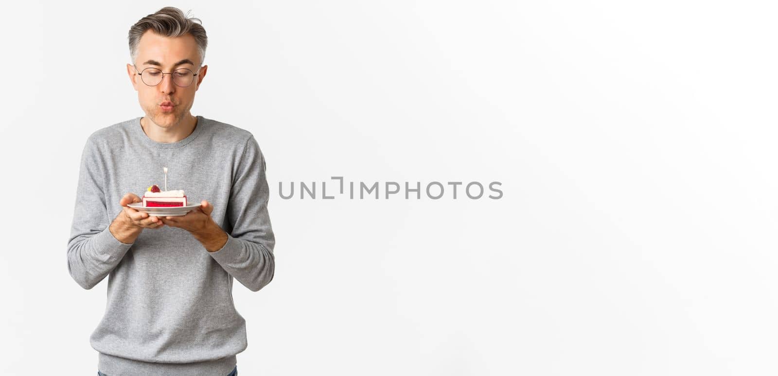 Portrait of attractive middle-aged man in grey sweater and glasses, celebrating his birthday, blowing a candle on b-day cake and making wish, standing over white background.