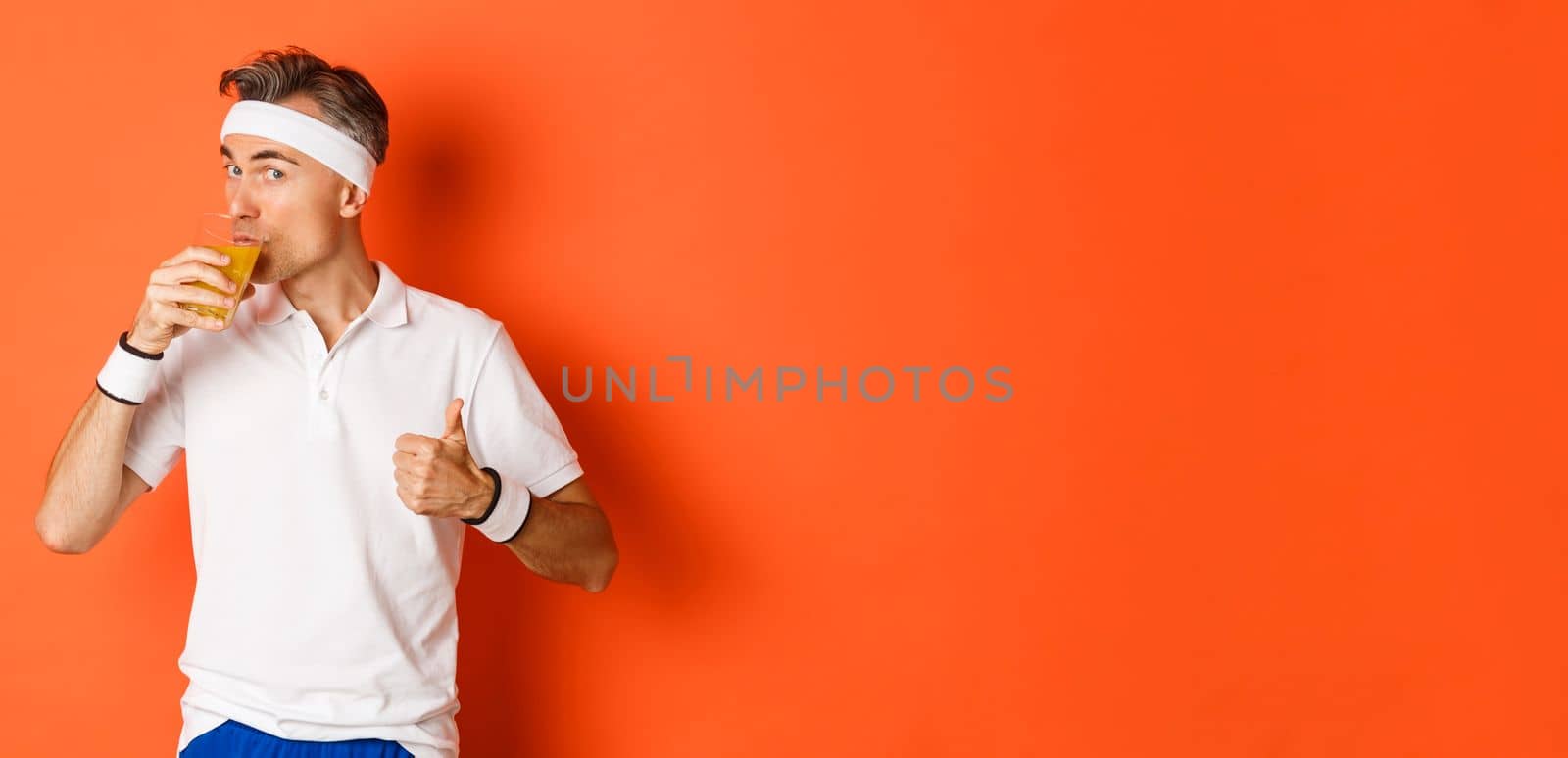 Concept of workout, gym and lifestyle. Portrait of handsome middle-aged fitness guy, showing thumbs-up and drinking juice, standing over orange background.
