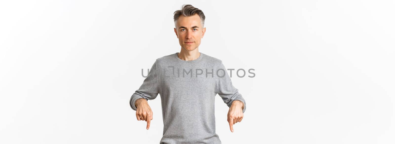 Portrait of successful middle-aged man in grey sweater, pointing fingers down and looking confident at camera, showing logo, standing over white background.