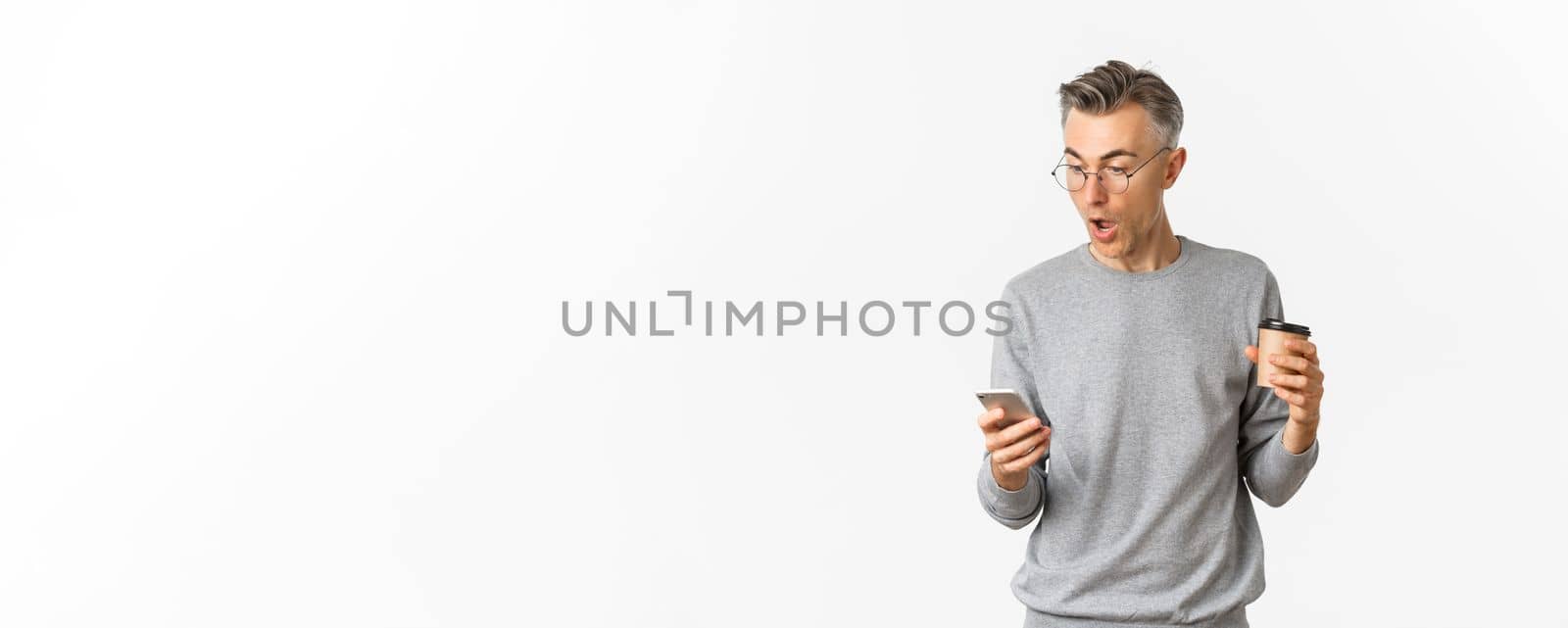 Image of handsome middle-aged man in grey sweater and glasses, drinking coffee and looking amazed at mobile phone screen, reading something online, standing over white background.