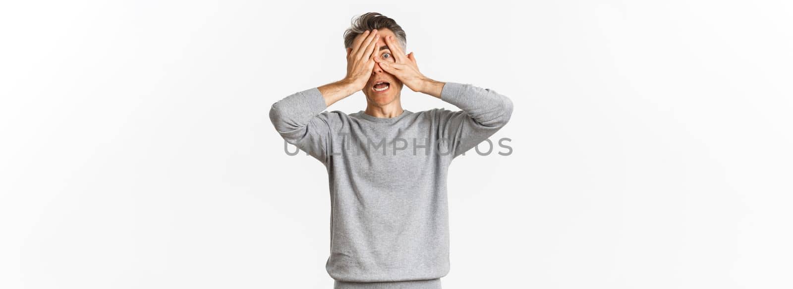 Image of embarrassed of scared middle-aged man cover his eyes with hands, peeking through fingers with shocked expression, standing over white background.