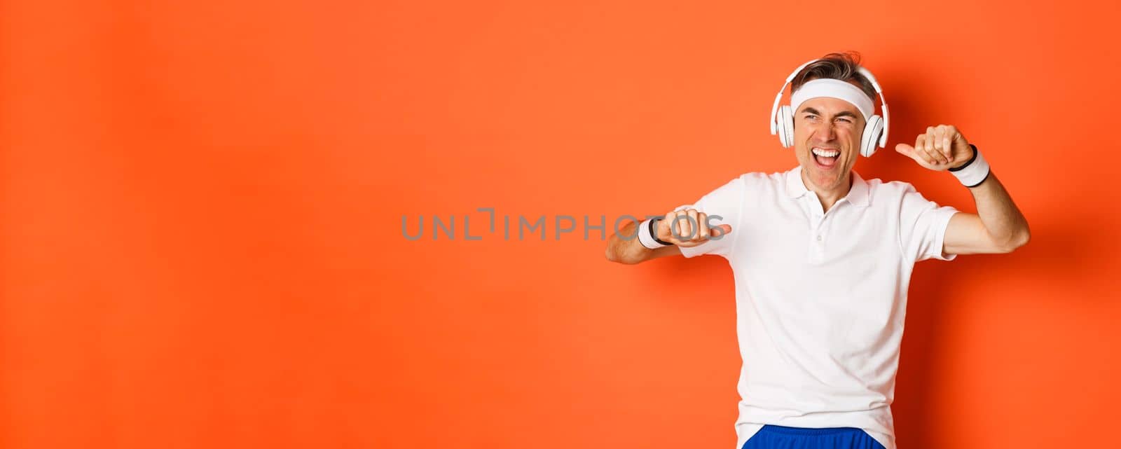 Portrait of handsome middle-aged male athlete, listening music in headphones during fitness training, dancing over orange background.