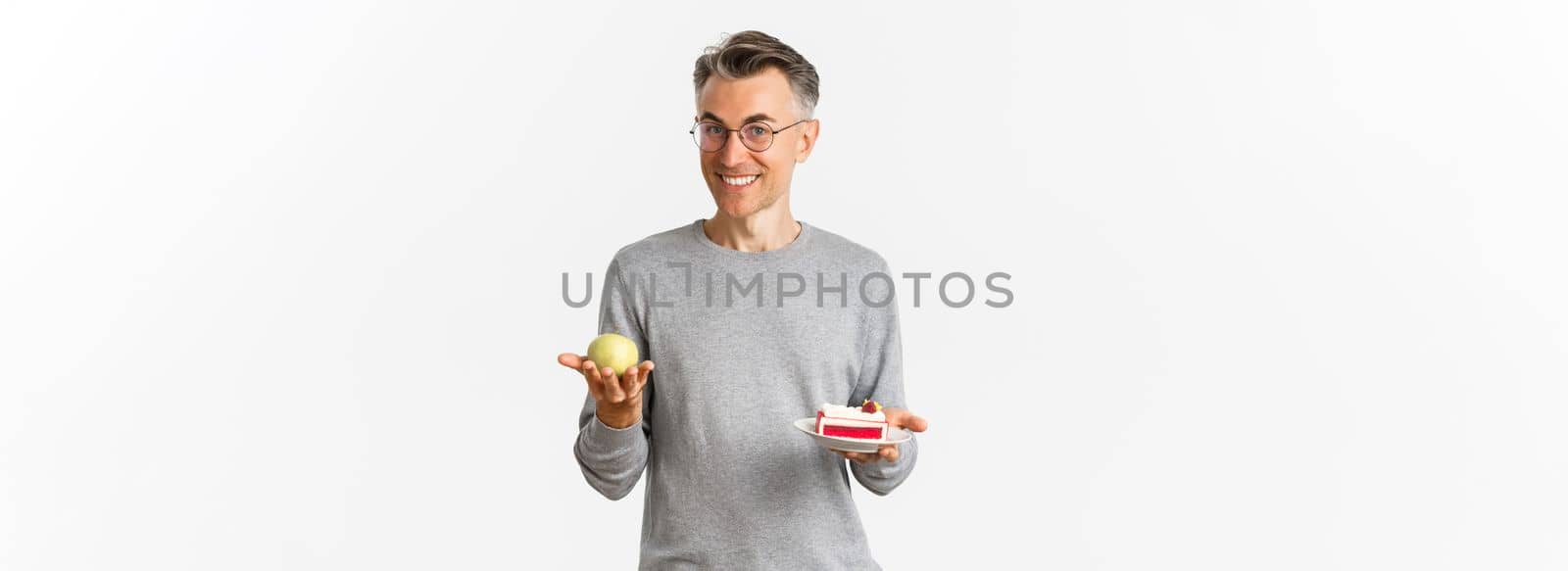 Image of handsome and healthy middle-aged man in glasses, recommending eat apple instead of cake, standing over white background.