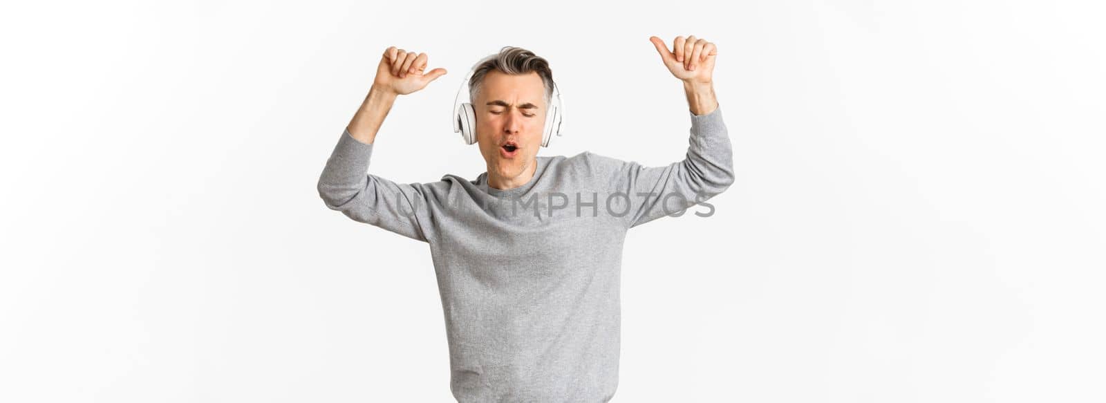 Portrait of handsome, modern middle-aged man, listening music in headphones and dancing, standing over white background.