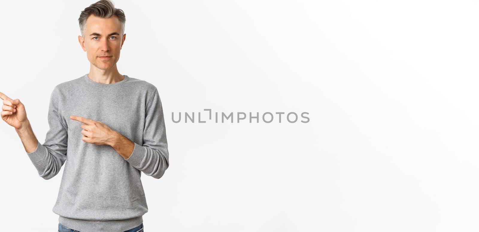 Portrait of handsome adult gay man in grey sweater, pointing fingers left and showing advertisement, standing over white background.