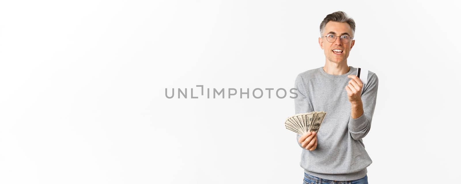 Portrait of excited middle-aged man looking amazed, holding money and showing credit card, standing over white background.