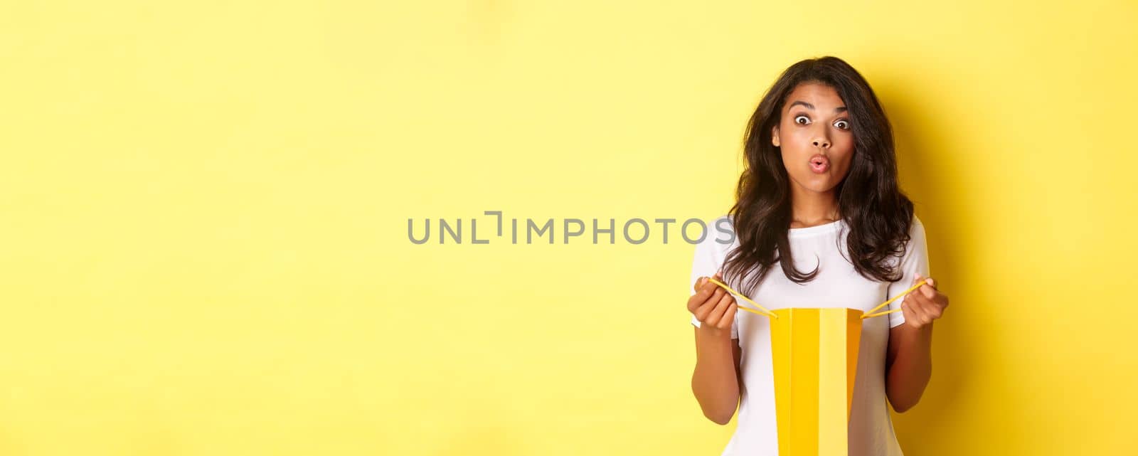 Image of surprised african-american girl receive a gift on holiday, open a shopping bag and looking amazed, standing over yellow background.