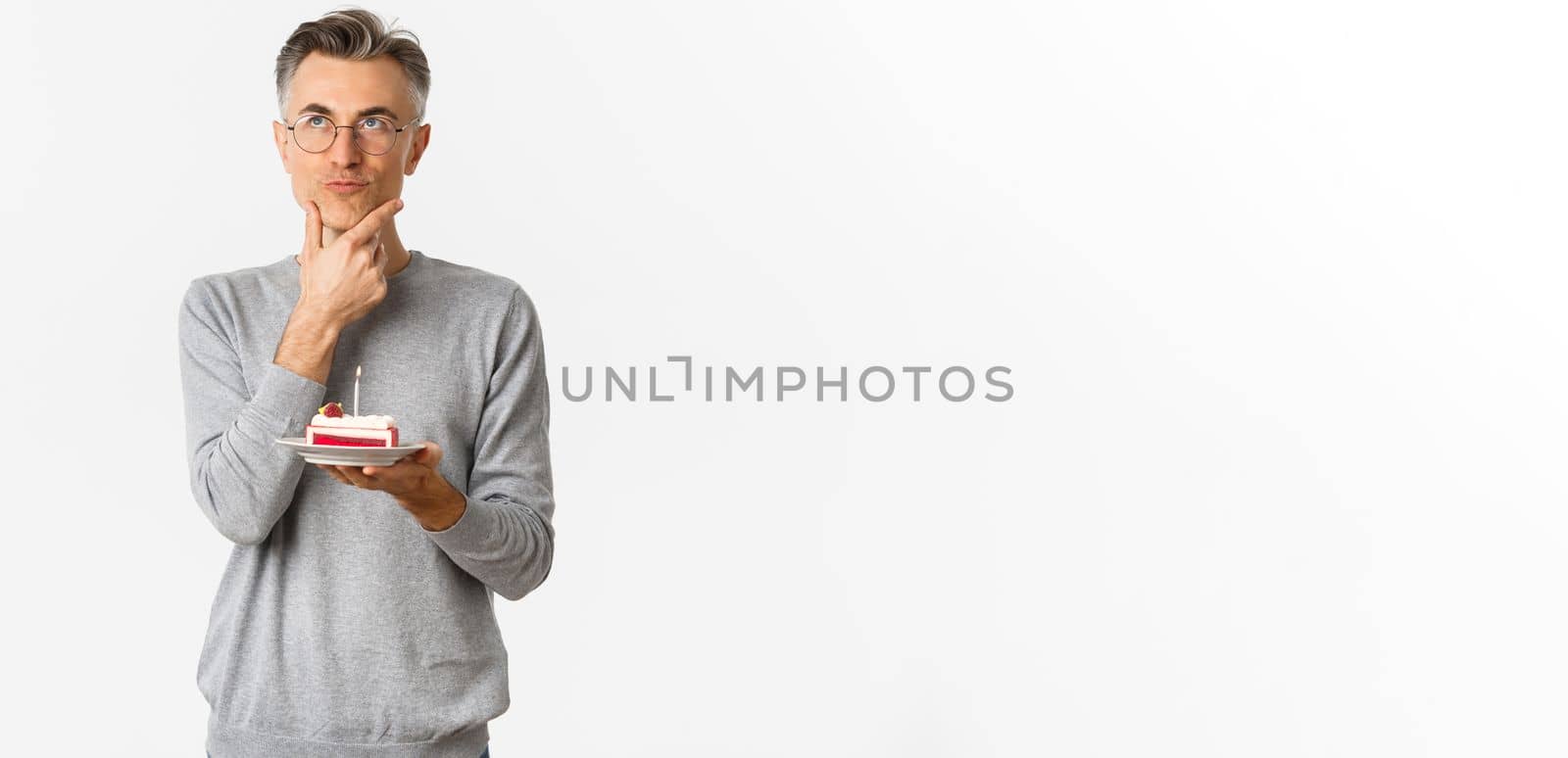Image of thoughtful and serious middle-aged man, celebrating birthday, holding b-day cake and thinking what wish to make, standing over white background.